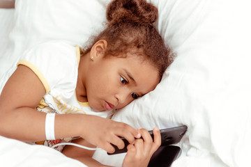 Cute little kid girl watching video on smartphone with smiley face alone on the bed, child using mobile phone with happy face at home. Stay at home quarantine coronavirus COVID-19 pandemic prevention.