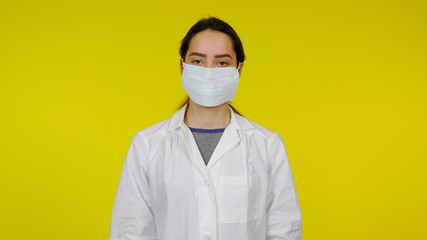 Young infectious disease doctor in a protective medical mask on her face looks at the camera. Girl in a white coat on a yellow background. Coronavirus, flu