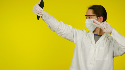 Young doctor in protective medical mask, glasses and latex gloves makes selfie on a smartphone. Nurse in a white coat on a yellow background with a phone in his hand