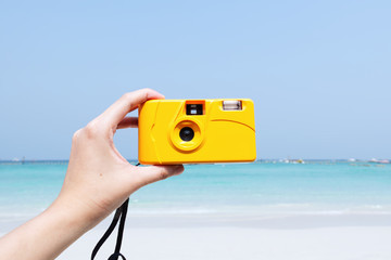 Hand holding yellow camera with beautiful summer beach background
