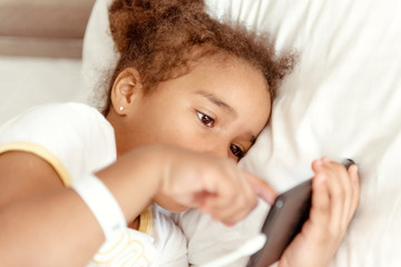 Cute little kid girl watching video on smartphone with smiley face alone on the bed, child using mobile phone with happy face at home. Stay at home quarantine coronavirus COVID-19 pandemic prevention.