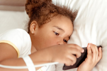 Cute little kid girl watching video on smartphone with smiley face alone on the bed, child using mobile phone with happy face at home. Stay at home quarantine coronavirus COVID-19 pandemic prevention.