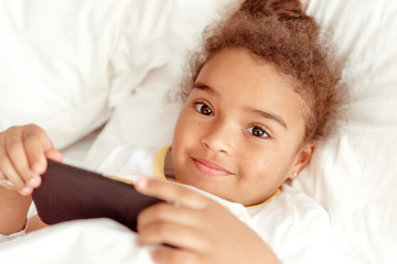 Cute little kid girl watching video on smartphone with smiley face alone on the bed, child using mobile phone with happy face at home. Stay at home quarantine coronavirus COVID-19 pandemic prevention.