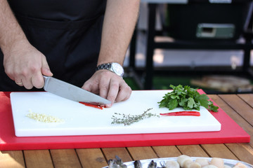 Preparation. Cooking process. The chef cuts red chili pepper with a knife on a white board on wooden table. Thyme,  coriander, herbs, chef hands. Background image, copy space