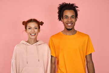 Positive dark skinned guy has date with ginger girlfriend, have fun together, pose at camera with broad smiles, pleased to rely on each other, hear something positive, make jokes together, pink wall