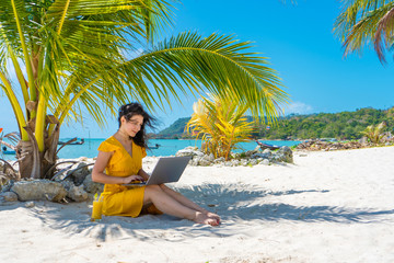 Fototapeta na wymiar Girl in a yellow dress on a tropical sandy beach works on a laptop and drinks fresh mango. Remote work, successful freelance. Works on vacation.