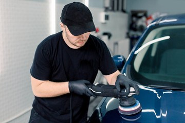Fototapeta na wymiar Car detailing and polishing concept. Professional Caucasian male car service worker, wearing black t-shirt and cap, holding in hands orbital polisher, and polishing blue luxury car in auto repair shop