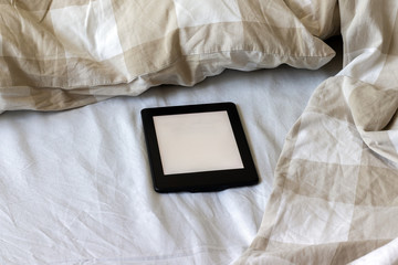 A modern black electronic book with a blank screen on a white and beige bed. Mockup tablet on bedding