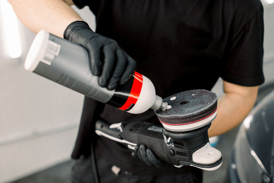 Cropped image of auto service worker, wearing black t-shirt and gloves, putting special polish wax or cream on the orbital polisher to polish the car behind