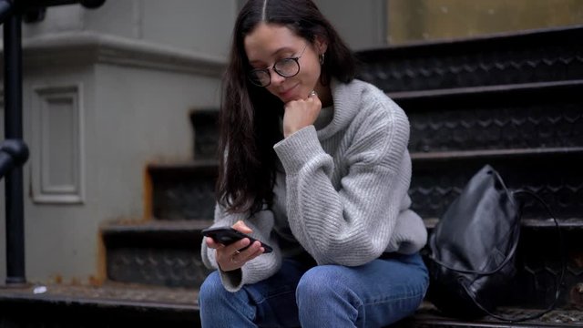 Caucasian female millennial in trendy outfit using 4g wireless for browsing internet and download media files on cellphone device, charming teenager sitting at city stairs and sending message 
