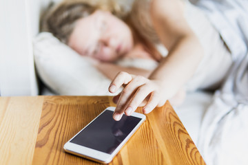 Young Eastern European woman lying in bed and turning off the alarm clock on her mobile phone in...
