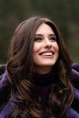 Girl smiling at camera on winter woods background. Glamorous funny young woman with smile wearing stylish sweater, purple fashionable fur coat. Fur and fashion concept. Beautiful people.