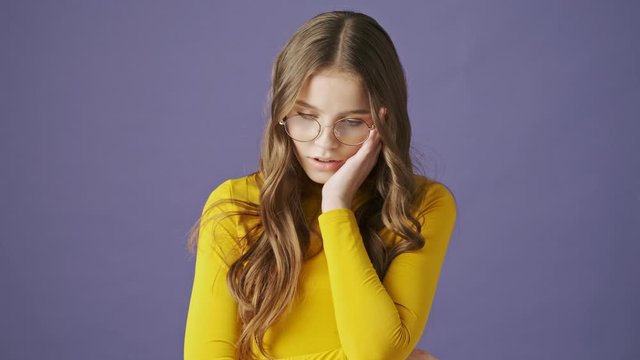 A frustrated teenage girl is touching her cheek while standing isolated over a purple background in studio