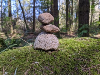 Fototapeta na wymiar A cairn of 3 small rocks set atop a large, mossy boulder, with trees and patches of sky in soft focus in the background. 