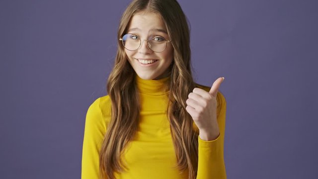 A beautiful smiling teenage girl is showing a thumb up standing isolated over a purple background in studio