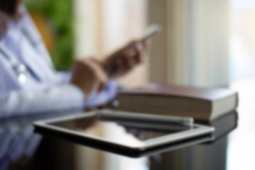 Blurred image of female doctor with  medical stethoscope hand using mobile smart phone with digital tablet computer and text book on the desk at office. Medical online networking, telehealth concept.