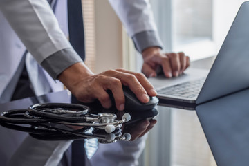 Male doctor hand using wireless mouse, browsing medical information on laptop computer with stethoscope on the desk. Medic tech, telehealth , online medical, teleconference or telemedicine concept. 