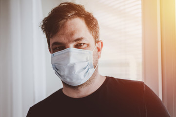 Man in quarantine during coronavirus COVID-19. Man with protective mask stands near window. Novel coronavirus 2019 COVID-19 theme. Quarantine during coronavirus pandemic