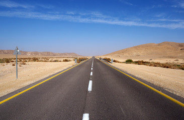 The desert landscape with the far yellow sandstone hills, the big empty black asphalt road, the blue sky with white clouds.