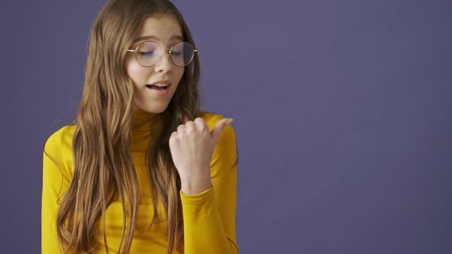 A happy positive teenage girl is looking to the side and then showing a thumb up standing isolated over a purple background in studio
