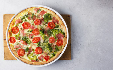 Open pie - quiche with bacon, broccoli, cherry tomatoes in white baking form on a wooden cutting board