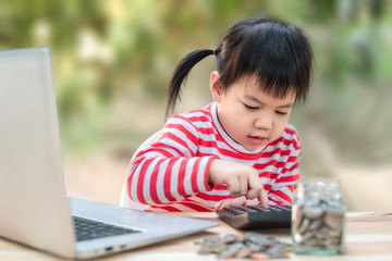 Fototapeta na wymiar Cute Asian little girl in red and white shirt, saving money for the future And accounting through a laptop and calculator. Money saving concept