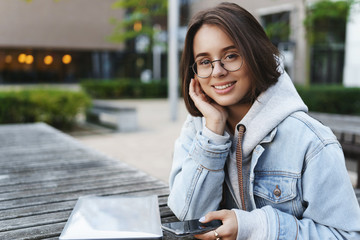 Close-up portrait of smiling attractive young woman in glasses and denim jacket, looking dreamy and...