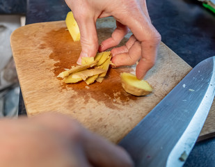 Human hand peeling fresh root ginger (Zingiber officinale) with a sharp kitchen knife on a wooden cutting board whilst preparing dinner, with selective focus