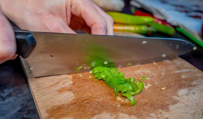 Close up of human hand chopping a hot green chilli pepper (Capsicum annuum) with a sharp kitchen knife on a wooden cutting board with selective focus on the chopped pepper