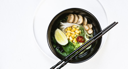 Asian cuisine vegetarian lunch. Flat-lay of Japanese rice noodle  soup with mushrooms, greens, corn, algae and chili peppers in black bowls on the white backgrond, top view, close up, wide composition
