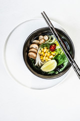 Asian cuisine vegetarian lunch. Flat-lay of Japanese rice noodle  soup with mushrooms, greens, corn, algae and chili peppers in black bowls on the white backgrond, top view