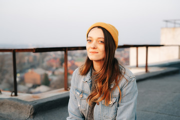 pretty white European woman is sitting on the roof of a multi-storey building in a blue denim jacket in gray pants,a yellow stylish hat.in the evening at sunset. lifestyle concept
