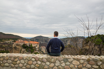 Naklejka na ściany i meble Man sits on a wall and looks at the Meteora monastery