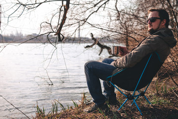 A man in sunglasses sits by the lake and looks into the distance. Portable folding, compact camping aluminum chair. Copy space, place for text