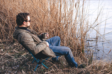 Naklejka na ściany i meble A man in sunglasses sits by the lake and looks into the distance. Portable folding, compact camping aluminum chair. Copy space, place for text