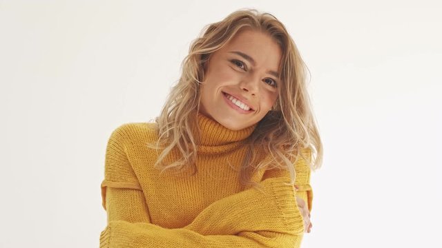 Pleased pretty blonde woman in sweater posing with crossed arms and looking at the camera over grey background