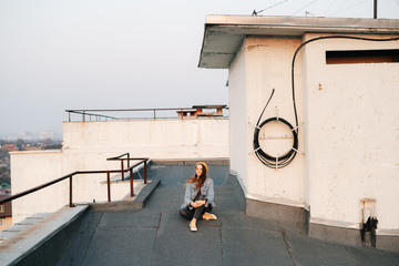 portrait pretty white European woman is sitting on the roof of a multi-storey building in a blue denim jacket in gray pants,a yellow stylish hat and yellow sneakers. lifestyle concept
