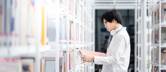 Smart Asian man university student wearing glasses choosing book from bookshelf. Textbook resources...
