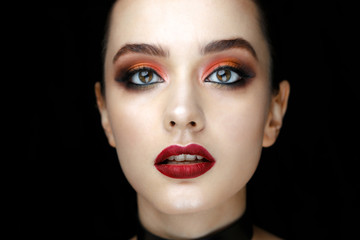 Close-up portrait of gorgeous young woman with red lips and bright makeup of eyes looks to the camera.. Beautiful girl with a healthy clean skin against black background.