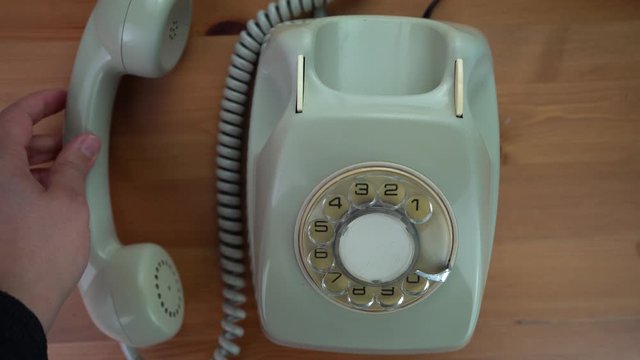 First-person shot. A hand picks up an antique white/green phone and leaves the handset on the table. Old fashioned. Zenithal shot of a classic phone. Subjective camera of an old style telephone dial. 