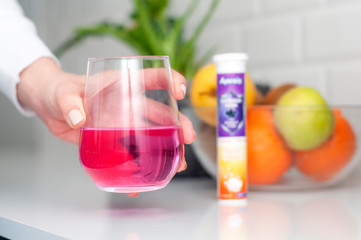 Caucasian woman holding glass of water with vitamin supplement in hand. Blurred effervescent tablet and fruit bowl on background. Female hand putting glass on white table. Boosting immune system.