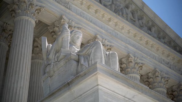 Slider to the right closeup of sculpture on the Supreme Court building of The Three Fates with Equal Justice slightly out of focus in the distance.