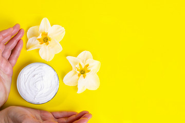 Human hands take cosmetic cream in jar and of blossoming flowers on bright yellow background. Skin care concept with natural creams. Top view. Flat lay. Copy space.