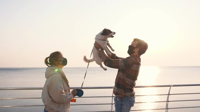 Young happy couple in protective medical masks playing with cute Jack Russel terrier dog outdoors near the sea during sunrise, slow motion