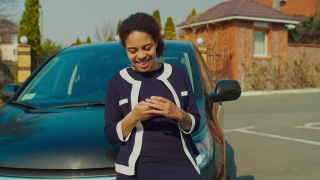 Cheerful happy african american woman in stylish clothes standing near brand new vehicle outdoors, holding car keys proudly, expressing joy and excitement after successful purchase in car dealership.