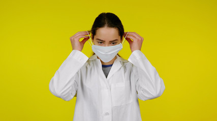 A young infectious disease doctor puts on a protective medical mask on her face. Girl in a white coat on a yellow background. Coronavirus, flu