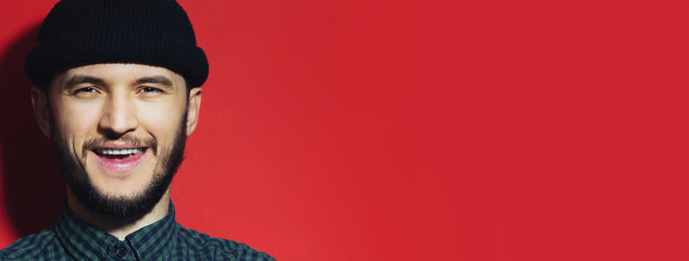 Smiling young guy wearing beanie black hat. Close-up portrait on red background with copy space.