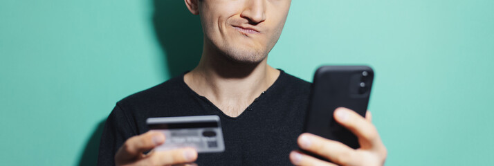 Panoramic close-up portrait of smiling guy holding smartphone and credit bank card on background of cyan, aqua menthe color.