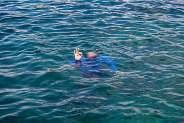 A man swimming underwater in the sea snorkeling, with a tube, in a blue suit, shows the OK sign