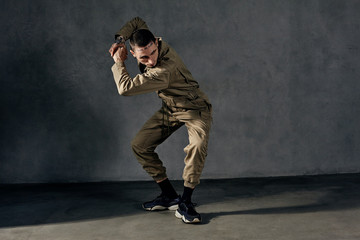 Fototapeta na wymiar Graceful guy with tattooed body and face, earrings, beard. Dressed in khaki overalls, black sneakers. Dancing on gray background. Dancehall, hip-hop